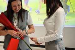 Dwie kobiety stoją naprzeciw siebie; jedna wręcza drugiej dokument, a obdarowana trzyma czerwoną różę i torebkę prezentową, uśmiechając się.