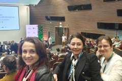 trzy kobiet siedzące na auli kongresu, obok i w tle inni uczestnicy, na ścianie ekran z wyświetloną prezentacją