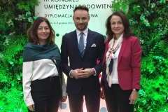 dwie kobiety i jeden mężczyzna stojący na tle ścianki promocyjnej oraz zieleni