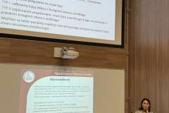 prelegent w sali konferencyjnej wygłasza wykład, na ścianie ekran z prezentacją