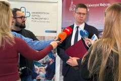 Mężczyzna w garniturze udziela wypowiedzi mediom; wokół niego stoją dziennikarze z mikrofonami, w tle baner Pomorskiego Uniwersytetu Medycznego