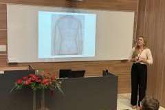 prelegent w sali konferencyjnej wygłasza wykład, na ścianie ekran z prezentacją