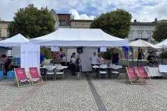 Grupa osób przy stoisku ucelnianym, pod namiotem ochronnym. Z przodu leżaki promocyjne uczelni