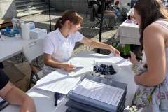 Młoda kobieta siedzi przy stoisku uczelni, tłumaczy coś osobie stojącej z drugiej strony stolika
