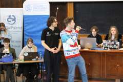 dwóch młodych mężczyzn stoi, jeden mówi przez mikrofon, z tyłu siedzą cztery młode kobiety