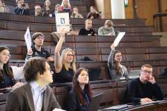 uczestnicy wydarzenia siedzący na auli konferencyjnej podnoszą białe kartki do góry