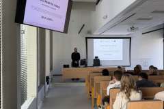 Prowadzący stoi przy katedrze, a studenci siedzą w ławkach i oglądają prezentację wyświetlaną na ekranie