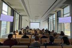 Prowadzący stoi przy katedrze, a studenci siedzą w ławkach i oglądają prezentację wyświetlaną na ekranie