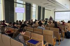 Prowadzący stoi przy katedrze, a studenci siedzą w ławkach i oglądają prezentację wyświetlaną na ekranie