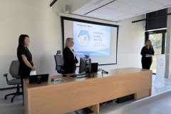 Trzy kobiety stoją przy biurku w sali wykładowej, obok ekranu z prezentacją.