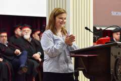 Młoda kobieta stoi przy mównicy i klaszcze z uśmiechem podczas uroczystości akademickiej; w tle siedzą profesorowie w togach i biretach