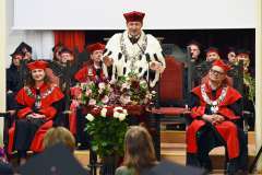 Rektor w białym gronostajowym płaszczu i czerwonym birecie stoi przy stole z kwiatami podczas uroczystości akademickiej