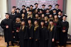 Grupa absolwentów w czarnych togach i biretach pozuje do wspólnego zdjęcia, trzymając czerwone dyplomy