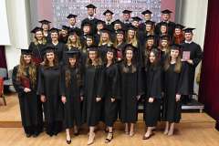 Grupa absolwentów w czarnych togach i biretach pozuje do wspólnego zdjęcia, trzymając czerwone dyplomy