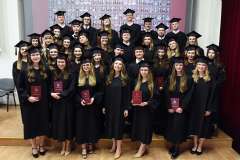 Grupa absolwentów w czarnych togach i biretach pozuje do wspólnego zdjęcia, trzymając czerwone dyplomy