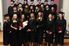 Grupa absolwentów w czarnych togach i biretach pozuje do wspólnego zdjęcia, trzymając czerwone dyplomy