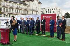 kobieta przemawia przy mównicy, w tle stoi grupa osób, ścianka reklamowa z logotypami oraz budynek szpitala