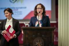 Kobieta przemawia przy mównicy, w tle stoi druga kobieta trzymająca dokumenty