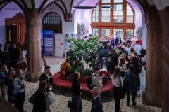 Hol budynku wypełniony uczestnikami wydarzenia; ludzie stoją i rozmawiają wokół czerwonej kanapy z roślinami, w tle widać wysokie okna