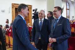 Trzech mężczyzn w garniturach rozmawia w sali audytoryjnej; w tle widoczna jest publiczność siedząca oraz kamera rejestrująca wydarzenie