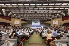 uczestnicy konwencji siedzą w sali audytoryjnej i słuchają wykładu, na ścianie wisi ekran wyświetlający prelegenta