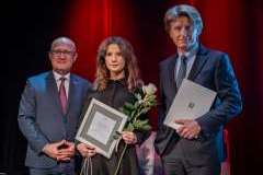 dwóch mężczyzn i kobieta stoją na scenie, kobieta trzyma różę i dyplom, jeden mężczyzna trzyma w ręku papierową teczkę