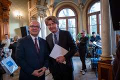 dwóch mężczyzn stojących w eleganckim wnętrzu, jeden trzyma w ręku papierową teczkę