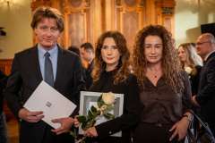 dwie kobiety i jeden mężczyzna stojący w eleganckich wnętrzach, jedna kobieta trzyma w ręku różę i dyplom, mężczyzna trzyma papierową teczkę