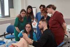 Grupa młodych osób w ubraniu medycznym w sali warsztatowej. na stole leżą fantomy do intubacji