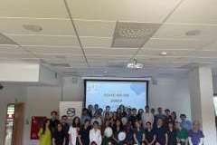 Grupa uczestników warsztatów w sali wykładowej, stojących przed ekranem rzutnika wyświetlającym napis NIME