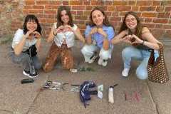 Cztery młode uśmiechnięte kobiety kucają na chodniku, dłonie ułożone w kształt serca. Przed nimi napis szczecin zrobiony z długopisów, parasolki, torb