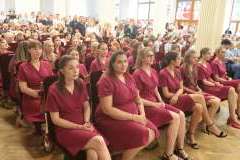 Grupa młodych osób w medycznych uniformach siedzi w sali z czerwonymi siedzeniami, w tle widoczne są duże okna i kolorowy witraż.