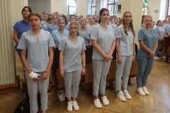 Grupa młodych osób w medycznych uniformach stoi w sali z czerwonymi siedzeniami, w tle widoczne są duże okna