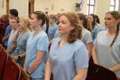 Grupa młodych osób w medycznych uniformach stoi w sali z czerwonymi siedzeniami, w tle widoczne są duże okna