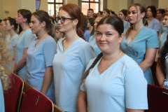 Grupa młodych osób w medycznych uniformach stoi w sali z czerwonymi siedzeniami, w tle widoczne są duże okna.