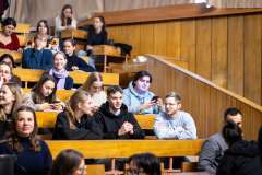 Uczestnicy siedzą w auli wykładowej na drewnianych ławkach, rozmawiają i przygotowują się do spotkania