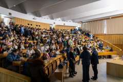 Pełna aula wykładowa z uczestnikami siedzącymi na ławkach oraz kilkoma osobami stojącymi przy mównicy przed rozpoczęciem spotkania