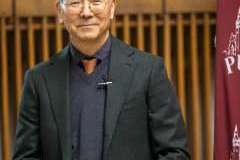 Mężczyzna w ciemnym garniturze i okularach stoi przed mównicą, przygotowany do wystąpienia