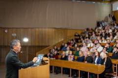 Mężczyzna w okularach przemawia przy mównicy, gestykulując ręką podczas wykładu