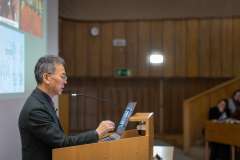 Mężczyzna w okularach przemawia przy mównicy, gestykulując ręką podczas wykładu