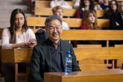 Mężczyzna w okularach siedzi w auli wykładowej, uśmiecha się, stoi przed nim butelka wody