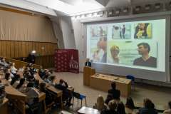 uczestnicy wykładu siedzący w auli wykładowej, przy mównicy stoi kobieta, w tle ekran ze slajdem
