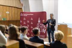 uczestnicy wykładu siedzący w auli wykładowej, na scenie stoi mężczyzna, gestykuluje, w tle slajd z prezentacji