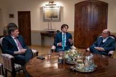 trzech mężczyzn w formalnych strojach siedzi przy stole i rozmawia