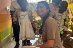 młoda kobieta w uniformie medycznym kuca i uśmiecha się. za nią siedzą dwie kobiety z dziećmi