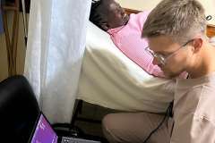 mężczyzna w uniformie medycznym siedzi przy łóżku szpitalnym na którym leży kobieta. kobieta uśmiecha się, medyk jest w trakcie robienia usg