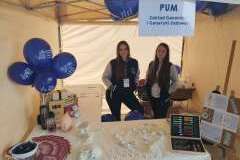 dwie kobiety pod namiotem ochronnym, na stoliku przed nimi modele kręgosłupa człowieka, czaszki, balony