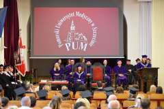 Uroczystość w auli – na scenie władze uczelni w fioletowych togach siedzą pod ekranem z logo PUM, a na widowni zgromadzeni są absolwenci i goście