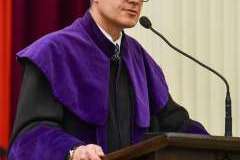 Mężczyzna w fioletowej todze i birecie przemawia przy drewnianej mównicy podczas uroczystości w auli