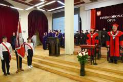 Uroczystość absolutoryjna w auli – poczet sztandarowy, władze uczelni w togach oraz przemówienie przy mównicy na tle napisu Finis coronat opus